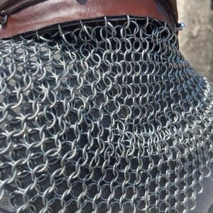 May include: Close-up of a chainmail coif with a brown leather neckband. The chainmail is made of small, interlocking metal rings. The leather neckband is secured with silver rivets.