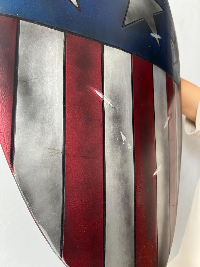 May include: A shield with a patriotic design, featuring red and white stripes and a blue field with a star. The shield has a curved shape and a distressed finish, with visible scratches and wear.