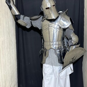 May include: A full suit of polished steel armor, including a helmet, chest plate, shoulder guards, and chainmail. The armor is worn with white pants and black gloves. A shield with a cross design is held in one hand.