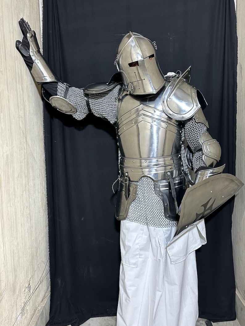 May include: A full suit of polished steel armor, including a helmet, chest plate, shoulder guards, and chainmail. The armor is worn with white pants and black gloves. A shield with a cross design is held in one hand.