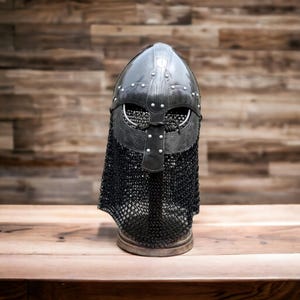 Può includere: Un elmo vichingo nero con una protezione per il collo a maglia di catena. L'elmo è in metallo e presenta rivetti e dettagli decorativi. L'elmo è su una superficie di legno contro uno sfondo di legno.