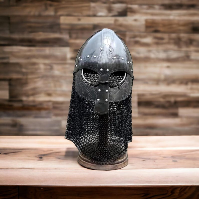 May include: A black Viking helmet with a chainmail neck guard. The helmet is made of metal and features rivets and decorative detailing. The helmet sits on a wooden surface against a wooden background.