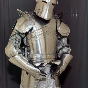 May include: A full suit of medieval armor made of polished steel. The armor includes a helmet with a cross design, shoulder guards, chest and back plates, arm and leg protection, and chainmail. The suit is designed to protect the wearer.