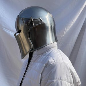Peut inclure: Un casque m&eacute;di&eacute;val argent&eacute; et poli, avec une visi&egrave;re et une cha&icirc;ne d&eacute;corative. Le casque est port&eacute; sur une veste blanche matelass&eacute;e, cr&eacute;ant une esth&eacute;tique historique et protectrice. La surface du casque refl&egrave;te la lumi&egrave;re ambiante.