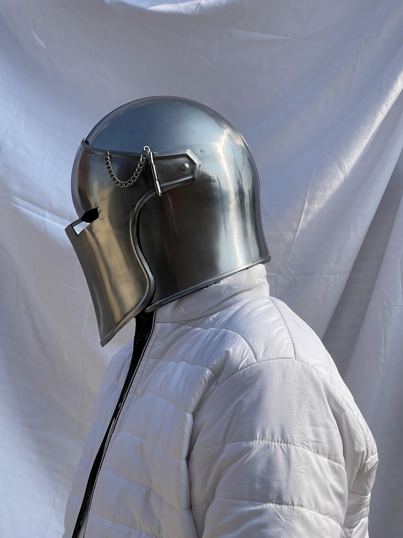 Peut inclure: Un casque m&eacute;di&eacute;val argent&eacute; et poli, avec une visi&egrave;re et une cha&icirc;ne d&eacute;corative. Le casque est port&eacute; sur une veste blanche matelass&eacute;e, cr&eacute;ant une esth&eacute;tique historique et protectrice. La surface du casque refl&egrave;te la lumi&egrave;re ambiante.