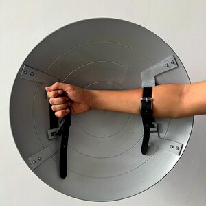 May include: A gray, circular shield with a series of concentric rings. The shield has black leather straps for securing to the arm. The shield is held by a hand. The shield is made of metal.