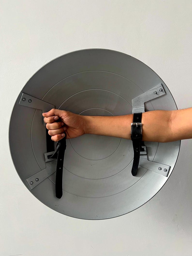 May include: A gray, circular shield with a series of concentric rings. The shield has black leather straps for securing to the arm. The shield is held by a hand. The shield is made of metal.