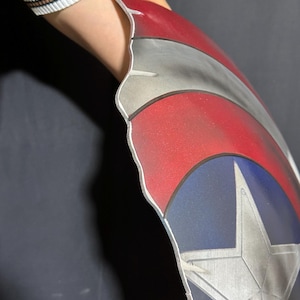 Op de afbeelding: Een Captain America schild rekwisiet, met een rood, wit en blauw kleurenschema en een zilveren ster embleem. Het schild heeft een gebogen vorm en is ontworpen om op de arm te worden gedragen. Het schild is gemaakt van een metalen materiaal.