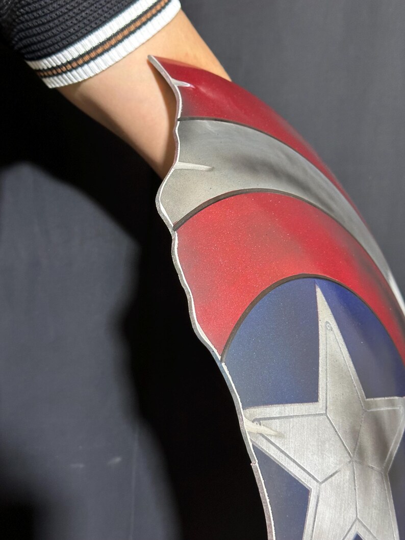 Op de afbeelding: Een Captain America schild rekwisiet, met een rood, wit en blauw kleurenschema en een zilveren ster embleem. Het schild heeft een gebogen vorm en is ontworpen om op de arm te worden gedragen. Het schild is gemaakt van een metalen materiaal.