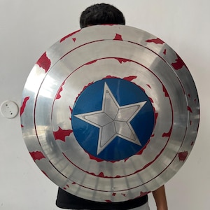 May include: A circular shield in silver and red, featuring a blue central star design. The shield appears to be a prop or costume piece, with a distressed finish for added effect.