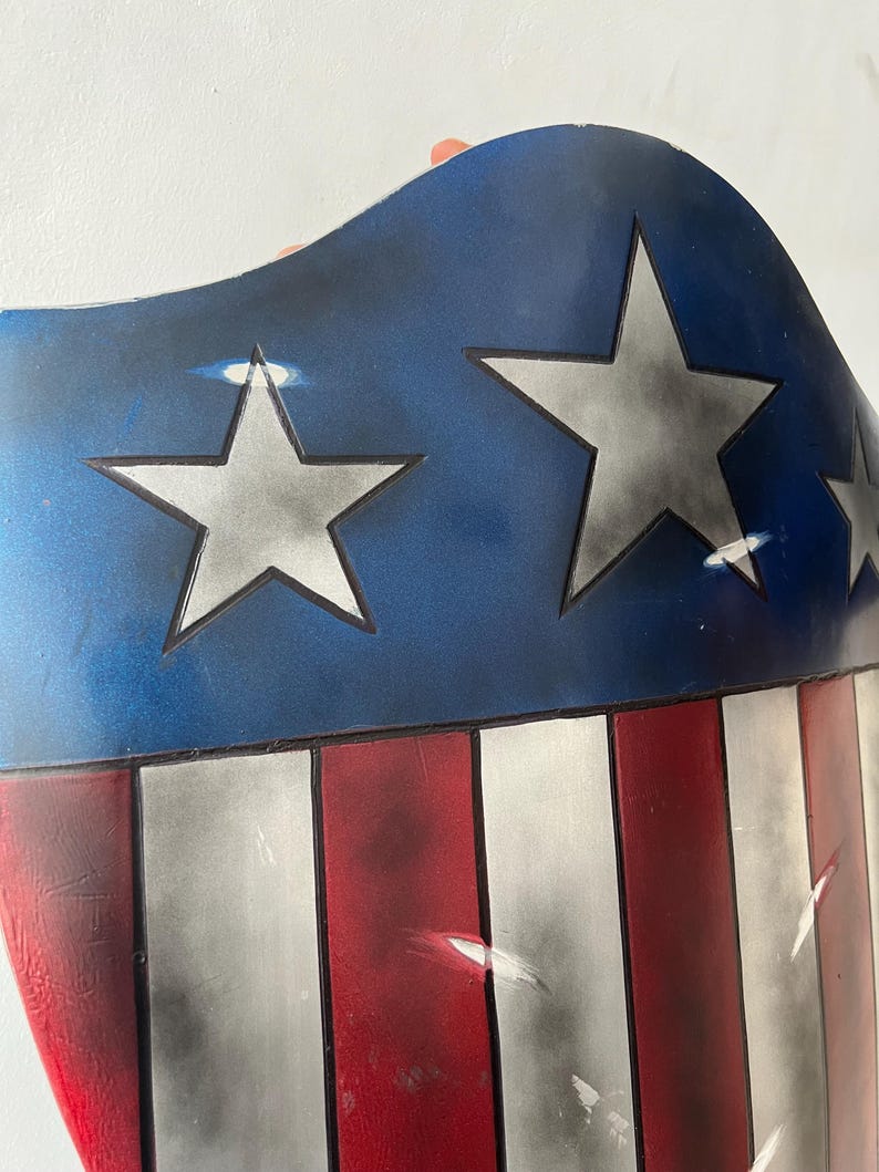 May include: A shield with a blue top section featuring white stars and a red and white striped bottom. The shield has a curved shape and appears to be made of metal or a similar material. The design is reminiscent of the American flag.
