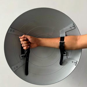 May include: A gray, circular shield with a series of concentric rings. The shield has black leather straps for securing to the arm. The shield is held by a hand. The shield is made of metal.