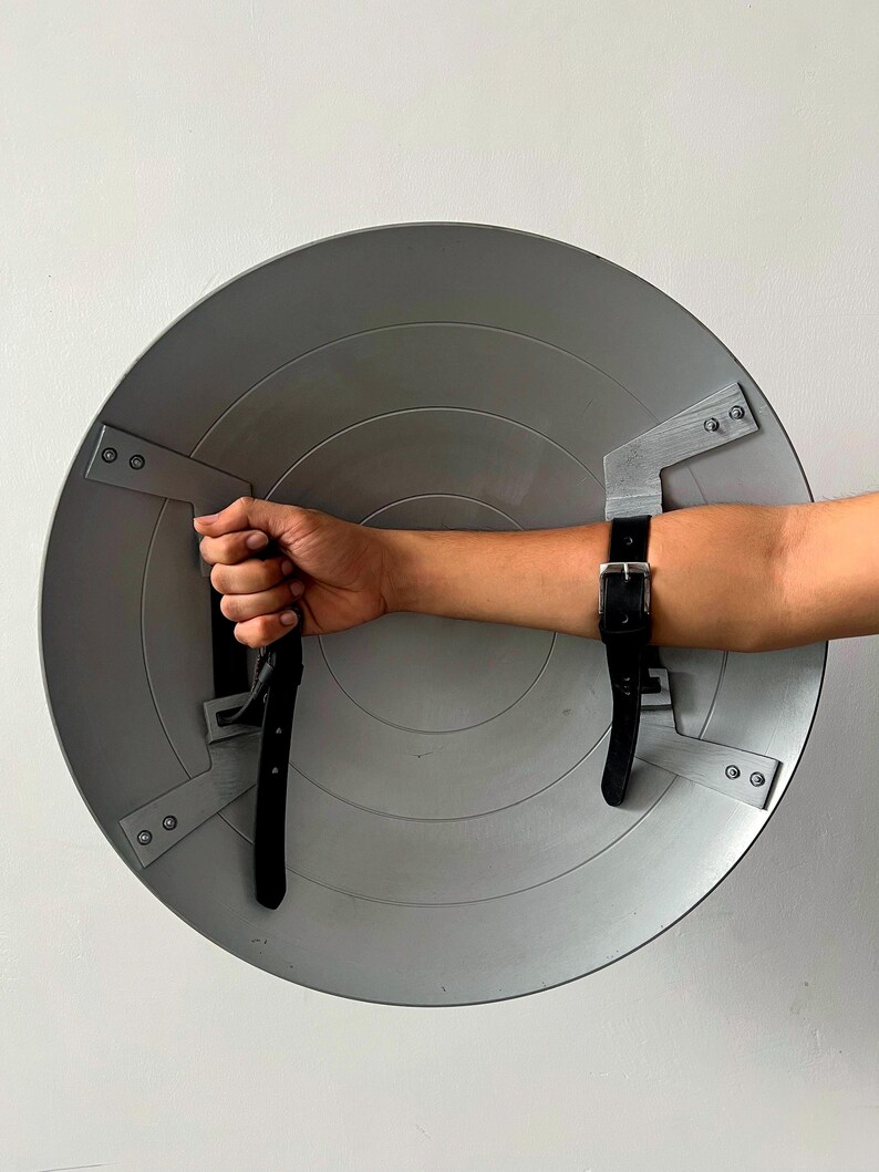 May include: A gray, circular shield with a series of concentric rings. The shield has black leather straps for securing to the arm. The shield is held by a hand. The shield is made of metal.