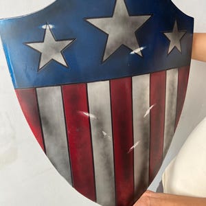 May include: A shield with a blue top section featuring three silver stars. The lower section has alternating red and white stripes. The shield is held up by a person.