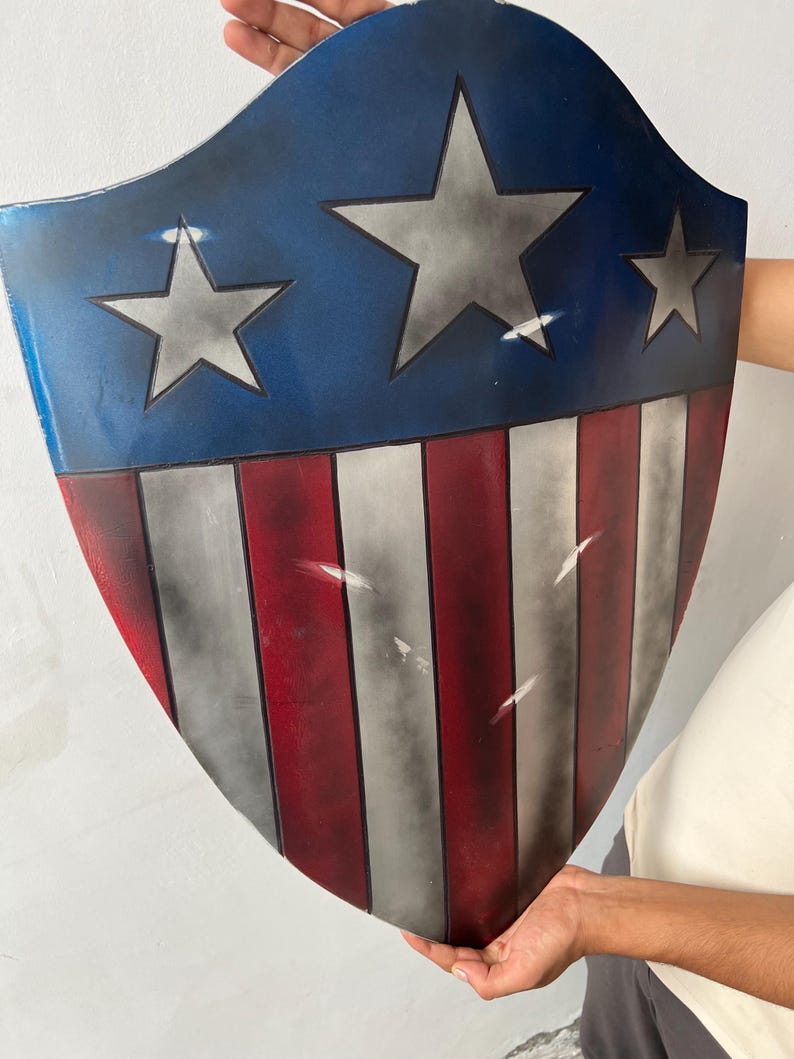 May include: A shield with a blue top section featuring three silver stars. The lower section has alternating red and white stripes. The shield is held up by a person.