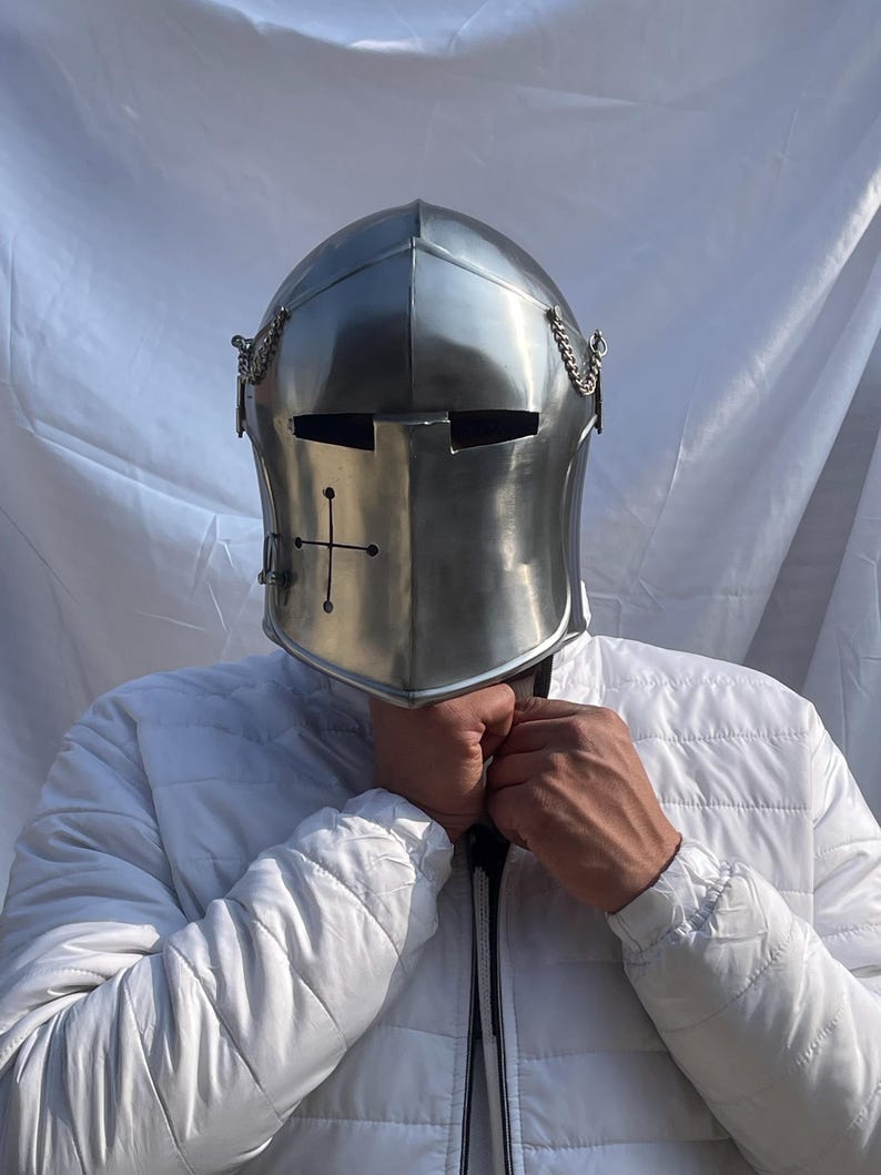 Peut inclure: Casque de chevalier argent&eacute; poli avec un embl&egrave;me de croix &agrave; l'avant. Le casque comporte des fentes pour les yeux et des d&eacute;tails de cha&icirc;ne. L'image montre le casque sur une personne portant une veste blanche.
