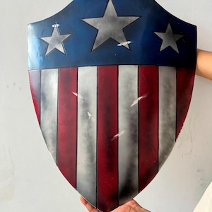 May include: A shield with a blue top section featuring three gray stars. The lower portion has red and white vertical stripes. The shield has a distressed appearance with visible wear and tear, suggesting a battle-worn aesthetic.