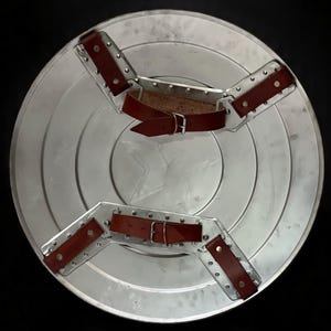 May include: A silver-colored, circular shield with a star emblem in the center. The shield has concentric rings and is equipped with two brown leather straps and metal brackets for securing it. The background is black.