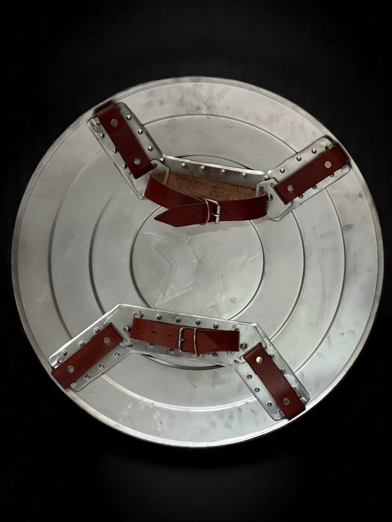 May include: A silver-colored, circular shield with a star emblem in the center. The shield has concentric rings and is equipped with two brown leather straps and metal brackets for securing it. The background is black.