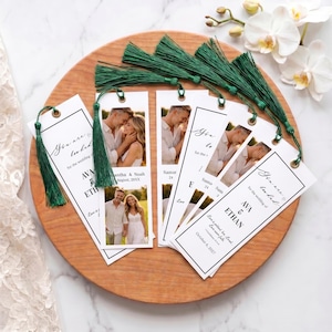 May include: A collection of white bookmarks with green tassels, arranged on a wooden tray. Each bookmark features a photo of a couple and text that reads "You are loved" and names. The bookmarks are for a wedding.