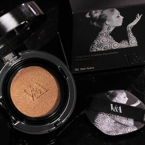May include: A black and silver compact with a gold-colored cushion foundation inside. The compact has the text "V&A" on it. The box for the compact is black with a white and black image of a woman and the text "Long-Wear Cushion Foundation SPF50+ PA+ 01 Fair Ivory V&A".