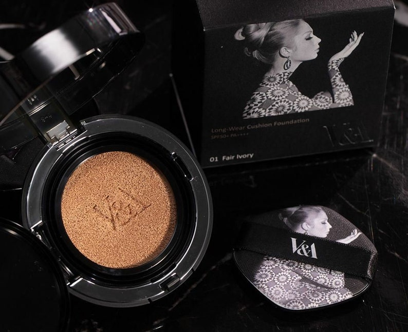 May include: A black and silver compact with a gold-colored cushion foundation inside. The compact has the text "V&A" on it. The box for the compact is black with a white and black image of a woman and the text "Long-Wear Cushion Foundation SPF50+ PA+ 01 Fair Ivory V&A".