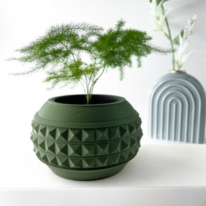 May include: A green ceramic planter with a geometric pattern, featuring a small green plant with long, thin leaves.