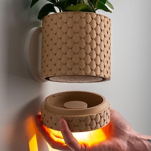 May include: A tan, textured, wall-mounted planter with a geometric pattern, holding a green plant. The planter has a matching base held in a hand, with a warm light emanating from it. The planter is attached to a white wall.