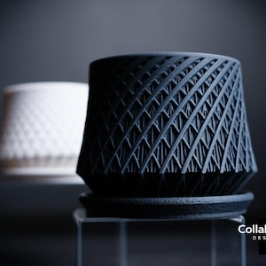 May include: Two contemporary plant pots are shown, one black and one white. The black pot features a geometric lattice design, while the white pot has a diamond pattern. The pots are likely for indoor plants and are displayed on a clear surface.