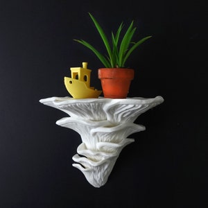 May include: A white, mushroom-shaped wall shelf with a yellow toy boat and a small potted plant. The plant has long green leaves and sits in an orange pot. The shelf is mounted on a dark wall.