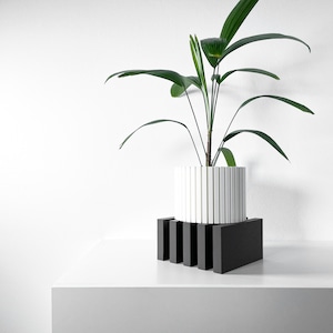 May include: A white planter with vertical lines sits on a black wooden stand. A green plant with long leaves grows from the planter.