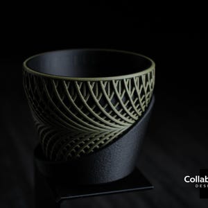 May include: A black and olive green decorative bowl with a leaf-like pattern. The bowl sits on a black stand. The interior of the bowl is black. The design is modern and minimalist.
