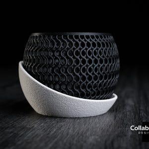 May include: A black, patterned, spherical planter sits on a white, textured saucer. The planter has a repeating, wave-like design. The design is a modern, decorative item for home or office decor.