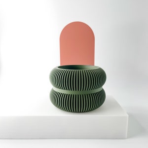 May include: A green, ribbed, double-tiered planter sits on a white platform in front of a pink, rounded shape.