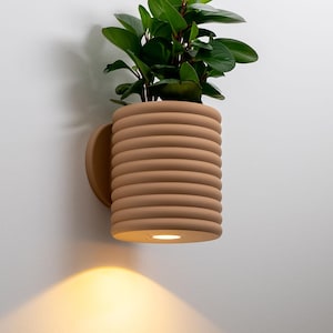 May include: A light brown wall-mounted planter with a ribbed design and a circular base. The planter holds a green plant. A small light is built into the base, shining downwards. The planter is mounted on a white wall.