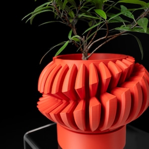May include: A bright orange, 3D-printed plant pot with a distinctive geometric pattern. The pot has a series of vertical, angular ridges around its spherical shape. A small green plant with slender leaves grows from the top, contrasting with the orange.