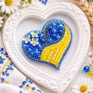Peut inclure: Une broche en forme de cœur ornée de perles bleues et jaunes. Le motif intègre de petites fleurs blanches, des perles bleues à facettes et des rangées de perles de rocaille jaunes. La broche est placée dans un cadre blanc en forme de cœur.
