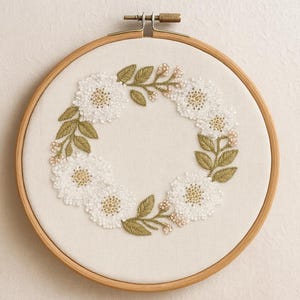 May include: Embroidered floral wreath on a wooden hoop. The wreath features white flowers with gold centres, green leaves, and small pink accents. The embroidery is set against a cream-coloured fabric, creating a delicate and handcrafted aesthetic.