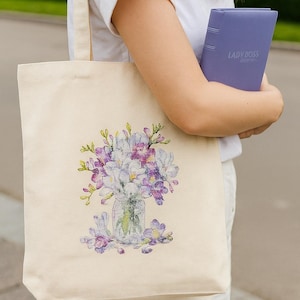 May include: Cream-colored tote bag featuring a floral design in shades of purple, white, and green. The bag is held by a person wearing a white shirt, along with a purple notebook labeled "LADYBOSS".