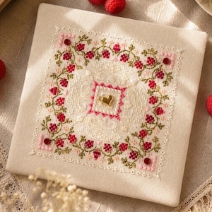 Könnte beinhalten: Ein quadratisches Stoffstück mit einer detaillierten Kreuzstichstickerei aus roten Beeren und grünen Ranken auf cremefarbenem Grund. Das Design enthält einen zentralen goldfarbenen Herzanhänger und vier kleine rote runde Verzierungen an den Ecken.