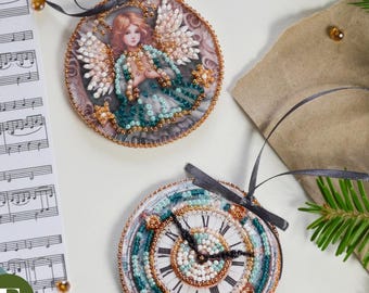 May include: Two round Christmas ornaments. One features an angel with wings, the other a clock face. Both are embellished with beads and have black ribbon hangers. A sheet of music and pine branches are in the background.
