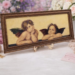 May include: A framed cross-stitch artwork featuring two cherubs with detailed wings and expressions. The artwork is set against a light yellow background and framed in a dark brown, ornate frame. The piece is displayed on a wooden easel.