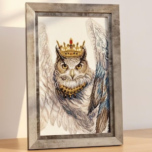 May include: Framed artwork featuring a detailed owl illustration. The owl is adorned with a golden crown and collar, set against a backdrop of detailed wings. The frame is a muted gray, adding a touch of elegance to the artwork.