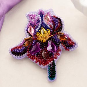 May include: A handcrafted iris flower brooch featuring intricate beadwork in shades of purple, red, and black. The design is accented with faceted crystals in pink, yellow, and deep purple, creating a textured, multi-dimensional floral appearance.