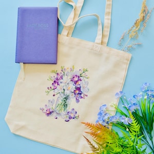 May include: A cream-colored tote bag with a floral design featuring purple and white flowers in a glass jar. A purple book with the text "LADY BOSS" is next to the bag. Artificial flowers and greenery are arranged around the bag on a blue surface.
