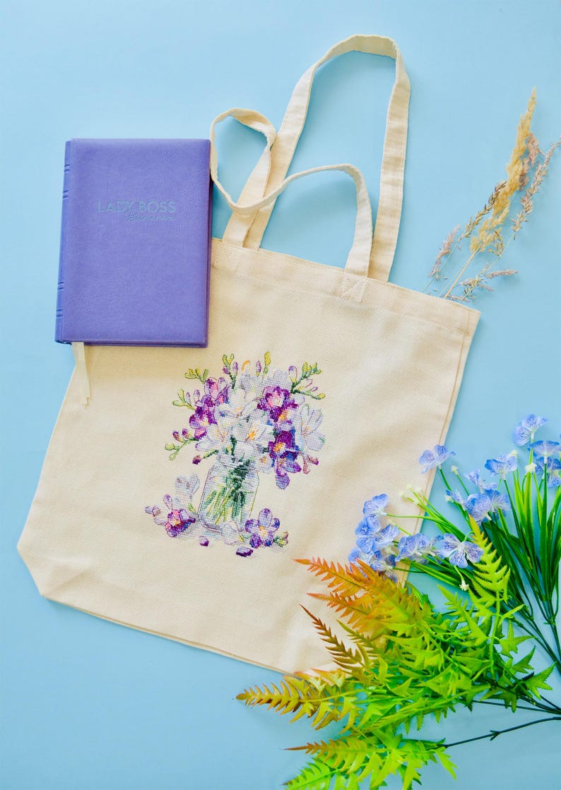 May include: A cream-colored tote bag with a floral design featuring purple and white flowers in a glass jar. A purple book with the text "LADY BOSS" is next to the bag. Artificial flowers and greenery are arranged around the bag on a blue surface.