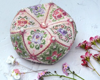 May include: A round, stuffed pincushion featuring intricate cross-stitch floral patterns in shades of pink, green, and purple, accented with small beads and a central pink bead. Two decorative pins with teapot-shaped heads are placed nearby.