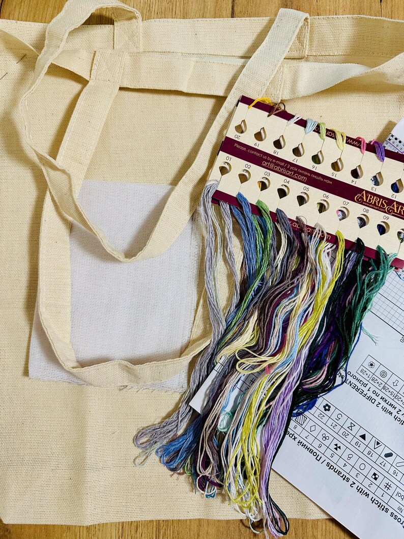 May include: A beige canvas tote bag with long handles, a white square pocket, and a colorful embroidery floss kit. The floss includes shades of blue, green, purple, and yellow. A chart with numbers and symbols is also visible.