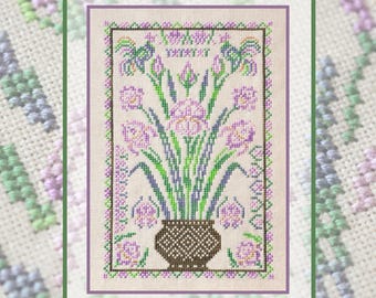 May include: A cross-stitch embroidery pattern on light beige fabric featuring a brown woven-style vase filled with purple and green iris flowers. The design is enclosed within a decorative border of small purple and green geometric shapes.