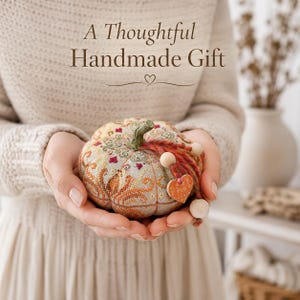 Finished pumpkin pincushion held gently in both hands as a thoughtful handmade gift. The decorative sampler features ceramic accents, wooden beads, decorative thread, and intricate embroidery. Ideal as a gift for stitchers, embroidery lovers, and fans of autumn handmade décor.